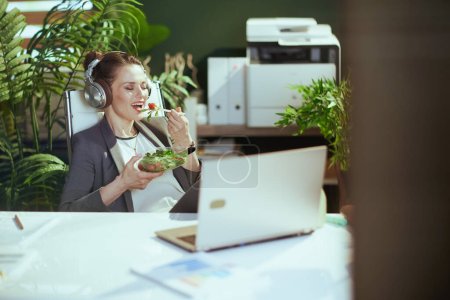 Téléchargez les photos : Un lieu de travail durable. employée moderne souriante dans un costume d'affaires gris dans un bureau vert moderne avec ordinateur portable et écouteurs manger de la salade. - en image libre de droit