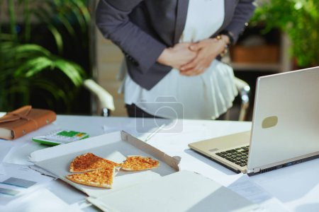 Foto de Lugar de trabajo sostenible. Primer plano de la mujer trabajadora en la oficina verde con pizza y portátil que tiene dolor de estómago. - Imagen libre de derechos