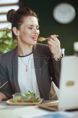 Foto de Lugar de trabajo sostenible. feliz mujer contable moderna de mediana edad en un traje de negocios gris en la oficina verde moderna con portátil comer ensalada. - Imagen libre de derechos