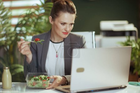 Téléchargez les photos : Un lieu de travail durable. moderne 40 ans propriétaire de petite entreprise femme dans un costume gris d'affaires dans le bureau vert moderne avec ordinateur portable manger salade. - en image libre de droit