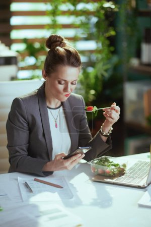 Photo for Sustainable workplace. sad modern 40 years old woman worker in a grey business suit in modern green office with laptop eating salad and using smartphone app. - Royalty Free Image