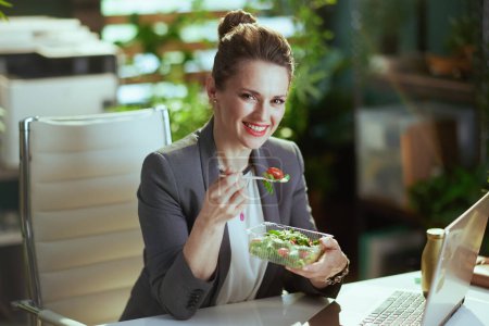 Téléchargez les photos : Un lieu de travail durable. heureuse femme d'âge moyen moderne travailleur dans un costume d'affaires gris dans un bureau vert moderne avec ordinateur portable manger salade. - en image libre de droit