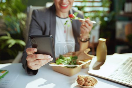 Photo for Sustainable workplace. happy modern middle aged small business owner woman in a grey business suit in modern green office with laptop eating salad and using smartphone app. - Royalty Free Image