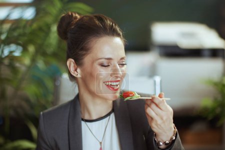 Téléchargez les photos : Un lieu de travail durable. souriant femme comptable d'âge moyen moderne dans un costume d'affaires gris dans le bureau vert moderne manger de la salade. - en image libre de droit