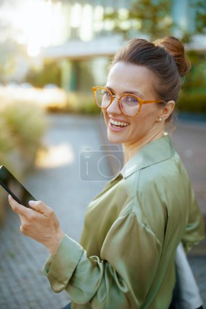 Téléchargez les photos : Souriant moderne employée dans le quartier des affaires en chemisier vert et lunettes à l'aide d'un smartphone. - en image libre de droit