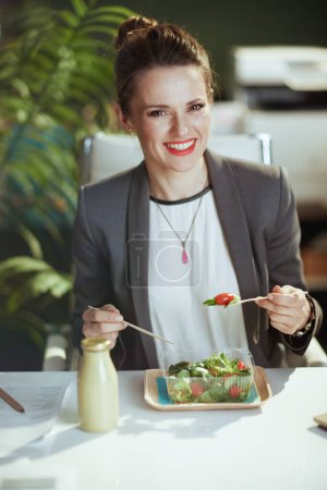 Foto de Lugar de trabajo sostenible. sonriente mujer contable moderna en un traje de negocios gris en la oficina verde moderna comer ensalada. - Imagen libre de derechos