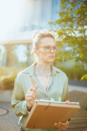 Photo for Pensive modern female worker near office building in green blouse and eyeglasses with documents and folder. - Royalty Free Image