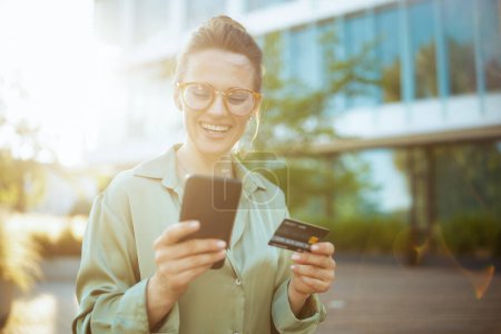 Foto de Sonriente trabajadora moderna en el distrito de negocios en blusa verde y gafas con tarjeta de crédito usando smartphone. - Imagen libre de derechos
