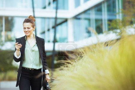 Téléchargez les photos : Heureuse employée moderne dans le quartier des affaires en veste noire en utilisant un smartphone et la marche. - en image libre de droit