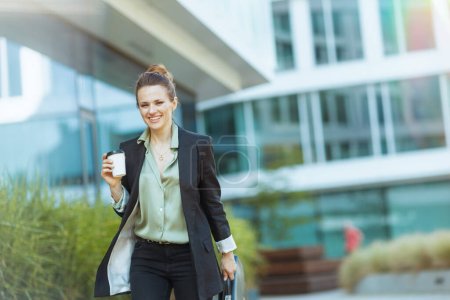Foto de Sonriente moderno 40 años de edad, empleada cerca de edificio de oficinas en chaqueta negra con taza de café caminando. - Imagen libre de derechos