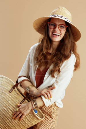 Téléchargez les photos : Vacances à la plage. souriant tendance 40 ans femme au foyer en chemisier blanc et short isolé sur fond beige dans des lunettes avec sac de paille et chapeau d'été. - en image libre de droit
