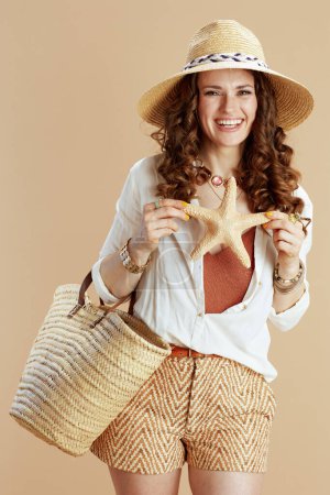 Photo for Beach vacation. happy elegant 40 years old woman in white blouse and shorts isolated on beige with sea star, straw bag and summer hat. - Royalty Free Image