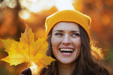 Photo for Hello september. smiling stylish female in brown coat and yellow hat with autumn yellow leaves outdoors in the city park in autumn. - Royalty Free Image