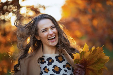 Téléchargez les photos : Bonjour, novembre. jeune femelle souriante en manteau beige avec des feuilles jaune automne à l'extérieur dans le parc de la ville en automne. - en image libre de droit