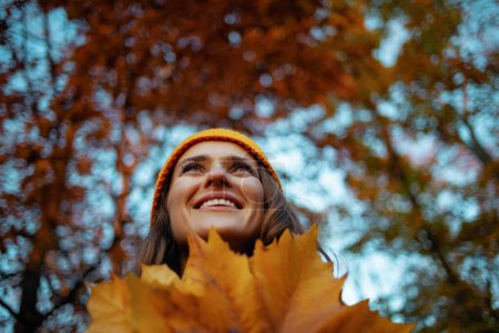 Foto de Hola otoño. sonriente mujer moderna en abrigo beige y sombrero naranja con hojas amarillas de otoño al aire libre en el parque de la ciudad en otoño. - Imagen libre de derechos