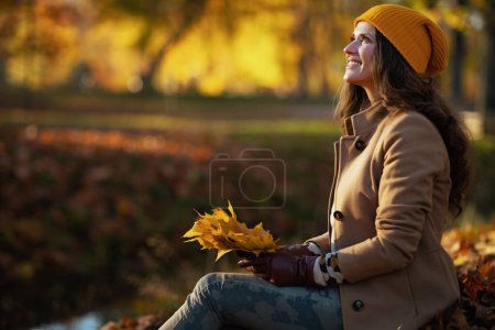 Foto de Hola septiembre. mujer moderna sonriente en abrigo marrón y sombrero amarillo con hojas amarillas de otoño sentado fuera en el parque de la ciudad en otoño. - Imagen libre de derechos