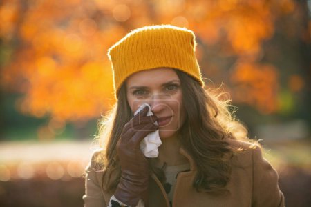 Photo for Hello november. trendy woman in brown coat and yellow hat with napkin blowing nose outside in the city park in autumn. - Royalty Free Image