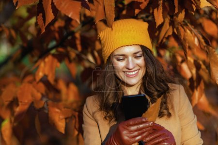 Hello november. smiling elegant woman in beige coat and orange hat using smartphone applications among autumnal foliage outside on the city park in autumn.