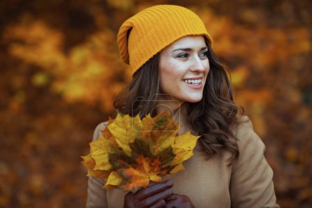 Photo for Hello november. happy modern female in beige coat and orange hat with autumn yellow leaves outside on the city park in autumn. - Royalty Free Image