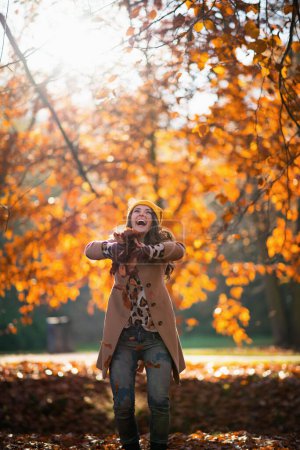 Photo for Hello september. happy trendy woman in beige coat and orange hat throws up autumn leaves outdoors on the city park in autumn. - Royalty Free Image