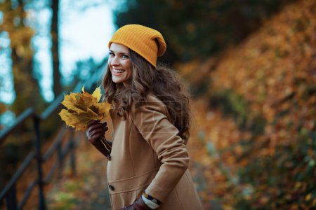 Photo for Hello autumn. happy stylish 40 years old woman in brown coat and yellow hat with autumn yellow leaves enjoying promenade outdoors in the city park in autumn. - Royalty Free Image