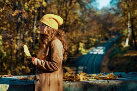 Foto de Hola otoño. mujer de mediana edad pensativa de moda en abrigo marrón y sombrero amarillo con hojas amarillas de otoño cerca del ferrocarril exterior en el parque de la ciudad en otoño. - Imagen libre de derechos