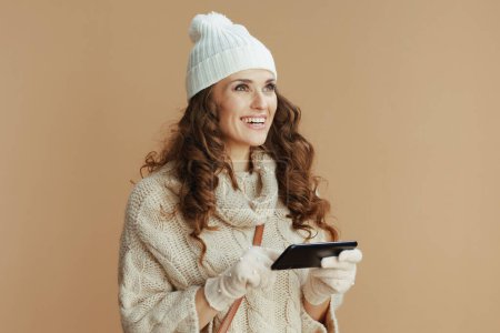 Photo for Hello winter. Portrait of happy elegant 40 years old woman in beige sweater, mittens and hat against beige background using smartphone applications. - Royalty Free Image