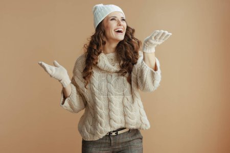 Téléchargez les photos : Bonjour l'hiver. Femme élégante souriante en pull beige, mitaines et chapeau isolés sur fond beige attrapant la neige. - en image libre de droit