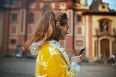 Foto de Sorprendida elegante turista solista en blusa amarilla y impermeable en Praga República Checa con teléfono inteligente. - Imagen libre de derechos