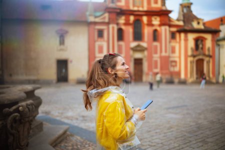 Téléchargez les photos : Femme moderne souriante en chemisier jaune et imperméable à Prague République tchèque avec smartphone marche. - en image libre de droit