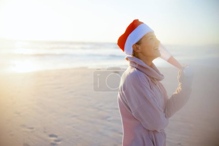 Photo for Happy modern 40 years old woman in cosy sweater with striped christmas hat at the beach in the evening. - Royalty Free Image