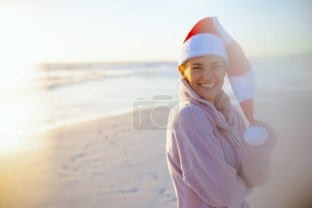 Photo for Portrait of happy modern middle aged woman in cosy sweater with striped christmas hat at the beach in the evening. - Royalty Free Image