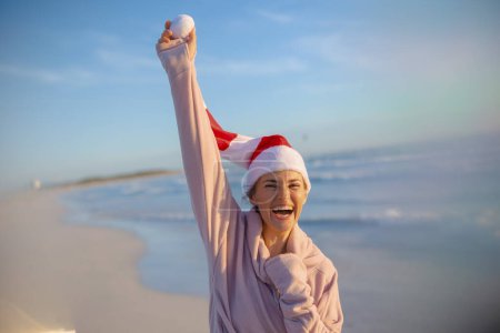 Foto de Sonriente mujer moderna en suéter acogedor con sombrero de Navidad a rayas regocijándose en la playa por la noche. - Imagen libre de derechos