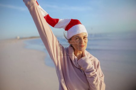 Photo for Happy modern woman in cosy sweater with striped christmas hat rejoicing at the beach in the evening. - Royalty Free Image