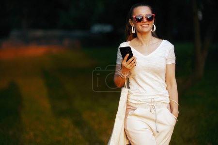 Téléchargez les photos : L'heure d'été. femme moderne souriante en chemise blanche en utilisant l'application smartphone et la marche. - en image libre de droit