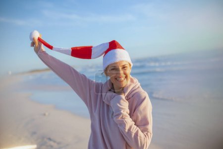 Photo for Smiling modern female in cosy sweater with striped christmas hat having fun time at the beach in the evening. - Royalty Free Image