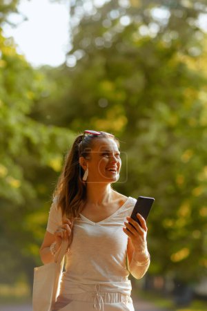 Foto de Hora de verano. mujer elegante sonriente en camisa blanca con bolso de mano usando teléfono inteligente y caminando en el parque de la ciudad. - Imagen libre de derechos