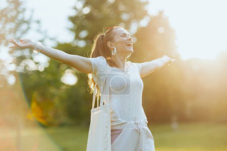 Foto de Hora de verano. sonriente mujer de 40 años en camisa blanca con bolso de mano regocijándose en el prado al aire libre. - Imagen libre de derechos