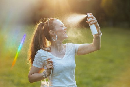 Téléchargez les photos : L'heure d'été. femme élégante souriante en chemise blanche en utilisant le spray visage dans la prairie à l'extérieur dans la nature. - en image libre de droit