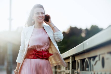 Photo for Happy stylish woman in pink dress and white jacket in the city speaking on a smartphone on the bridge. - Royalty Free Image
