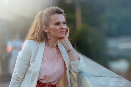 Photo for Pensive stylish woman in pink dress and white jacket in the city on the bridge. - Royalty Free Image