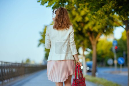 Foto de Visto desde atrás mujer en vestido rosa y chaqueta blanca en la ciudad escuchando la música con auriculares. - Imagen libre de derechos