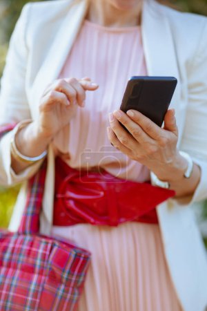Téléchargez les photos : Gros plan sur la femme en robe rose et veste blanche dans la ville avec smartphone et sac rouge. - en image libre de droit