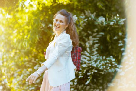 Téléchargez les photos : Sourire élégante femme de 40 ans en robe rose et veste blanche dans la ville contre la verdure. - en image libre de droit