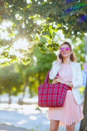 Foto de Mujer de moda feliz en vestido rosa y chaqueta blanca en la ciudad con bolsa roja y gafas de sol usando un teléfono inteligente. - Imagen libre de derechos