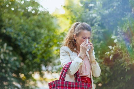 Téléchargez les photos : Jeune femme en robe rose et veste blanche dans la ville avec serviette et sac rouge a une attaque d'allergie. - en image libre de droit