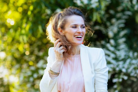 Photo for Happy modern woman in pink dress and white jacket in the city against greenery. - Royalty Free Image