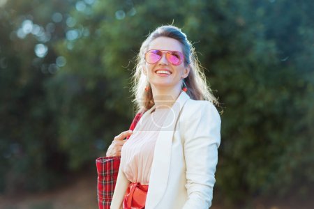 Téléchargez les photos : Jeune femme souriante en robe rose et veste blanche dans la ville avec sac rouge et lunettes de soleil. - en image libre de droit