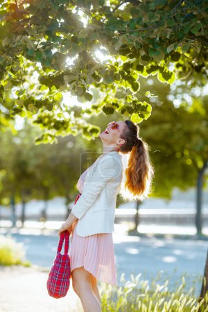 Téléchargez les photos : Heureuse femme élégante en robe rose et veste blanche dans la ville avec sac rouge et lunettes de soleil. - en image libre de droit