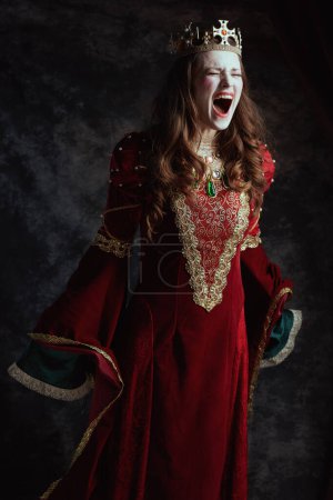 Photo for Mad medieval queen in red dress with white makeup and crown on dark gray background. - Royalty Free Image